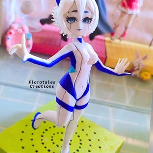 K&ouml;nnte beinhalten: Eine wei&szlig;e und blaue Figur einer weiblichen Figur mit Katzenohren. Die Figur ist mit einem erhobenen Bein und dem anderen gebeugten Bein posiert. Die Figur steht auf einem gr&uuml;nen Sockel mit L&ouml;chern.