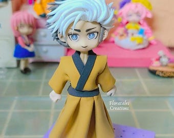 May include: A handmade figurine of a person with white and black hair wearing a yellow and black robe. The figurine is standing on a purple base with small holes.