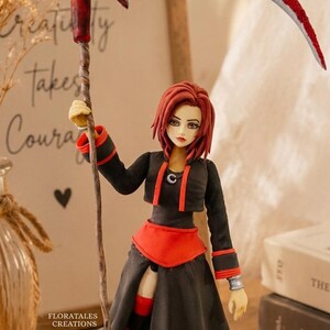 May include: A handcrafted figurine of a character with red hair, wearing a black and red outfit, and holding a red scythe. The figurine is standing in front of a framed print with the text "Creativity takes Courage". The figurine is a Floratales Creation.