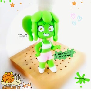 May include: A handmade green figurine of a cartoon character with a green head and hair, wearing a white top and pink shorts. The figurine is standing on a wooden base. The image includes the text "FLORATALES CREATIONS" and "SNAILED IT".