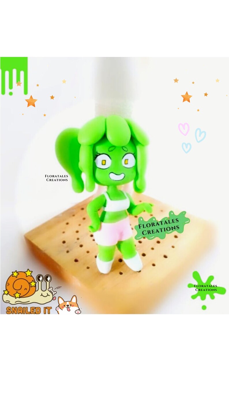 May include: A handmade green figurine of a cartoon character with a green head and hair, wearing a white top and pink shorts. The figurine is standing on a wooden base. The image includes the text "FLORATALES CREATIONS" and "SNAILED IT".