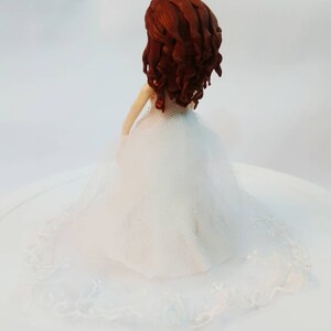 K&ouml;nnte beinhalten: Ein wei&szlig;er Hochzeitstortenaufsatz mit einer Brautfigur mit braunen Locken und einem wei&szlig;en Kleid mit Spitzenbesatz.