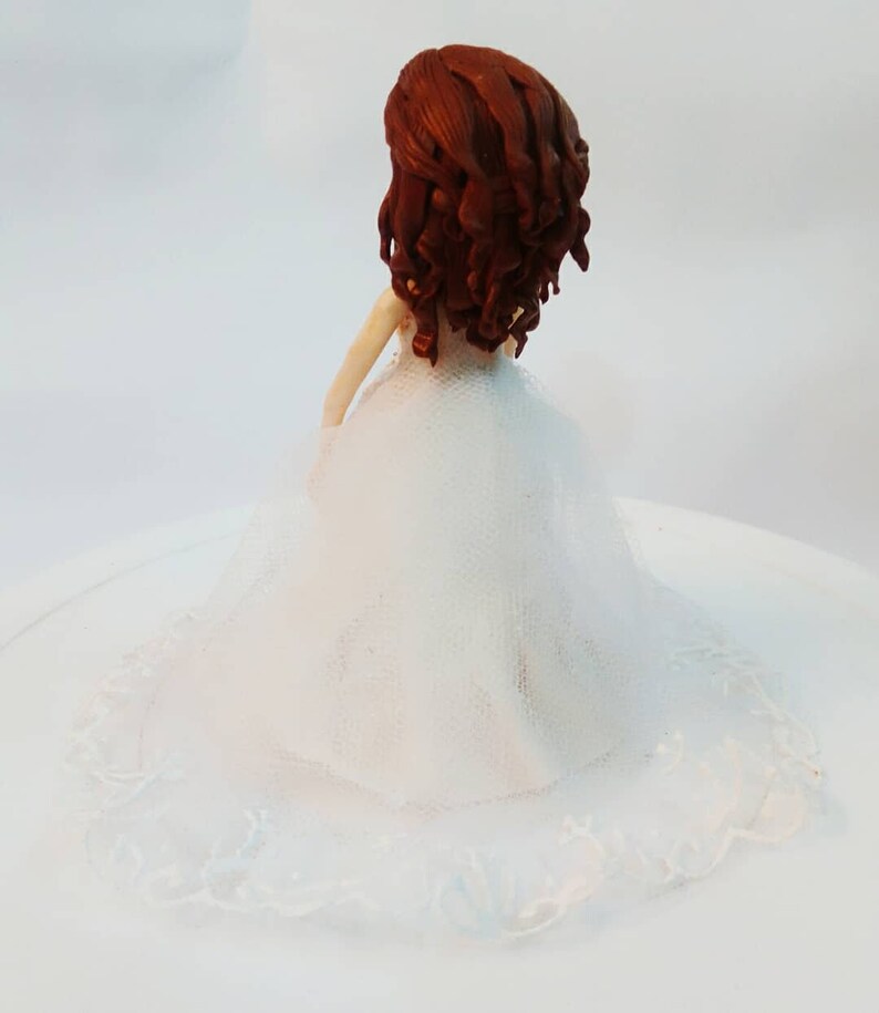 K&ouml;nnte beinhalten: Ein wei&szlig;er Hochzeitstortenaufsatz mit einer Brautfigur mit braunen Locken und einem wei&szlig;en Kleid mit Spitzenbesatz.