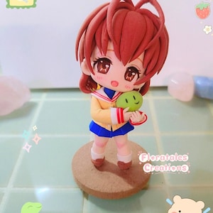 May include: A handcrafted figurine of an anime-style character with auburn hair, wearing a yellow sailor uniform with a blue skirt, holding a green creature. The figurine stands on a round, brown base. The text "Floratales Creations" is visible.