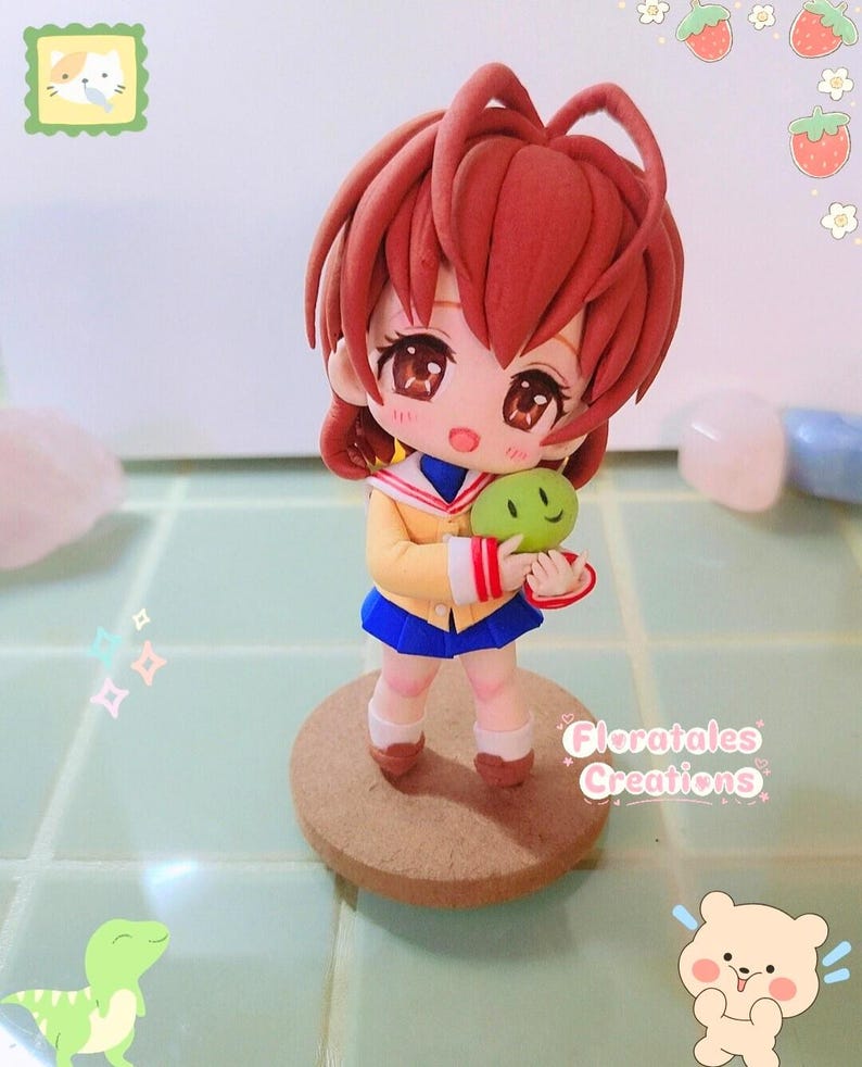 May include: A handcrafted figurine of an anime-style character with auburn hair, wearing a yellow sailor uniform with a blue skirt, holding a green creature. The figurine stands on a round, brown base. The text "Floratales Creations" is visible.