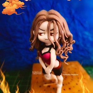 May include: A handcrafted figurine of a female character with long brown hair, red eyes, and a determined expression. She wears a pink and black outfit, posed with a fiery background. The figurine is a unique art piece, with the text "FLORATALES CREATIONS" visible.