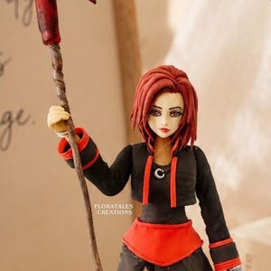 May include: A handcrafted figurine of a female anime character with red hair, wearing a black outfit with red accents. She holds a scythe with a red blade. The figurine is detailed with a stylized design. The text "FLORATALES CREATIONS" is visible.