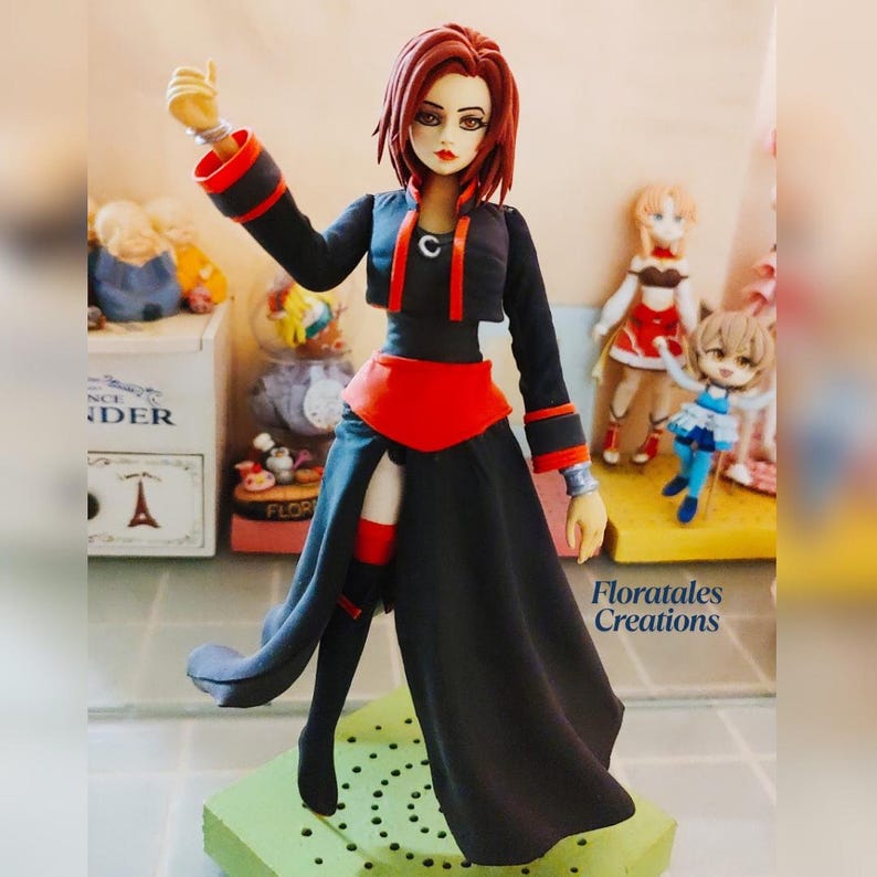 May include: A detailed figurine of a woman with red hair, wearing a black outfit with red accents. The figure is posed with one arm raised. The base is green, and the background includes other figurines and a decorative box.