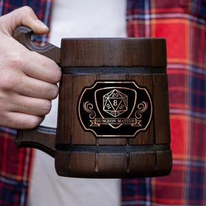 May include: A dark brown wooden beer stein with a handle, featuring a black and gold emblem with a D20 die and the words "Dungeon Master." The stein is held by a person wearing a red and blue plaid shirt.