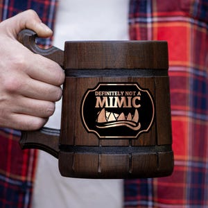 Puede incluir: Un jarro de cerveza de madera marrón oscuro con asa. El jarro tiene una placa decorativa con las palabras "DEFINITELY NOT A MIMIC" en blanco y dorado. La persona lleva una camisa a cuadros roja y negra.