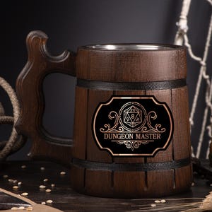 May include: A dark brown wooden beer mug with a carved handle and a metal interior. The mug features a black label with gold lettering that reads "DUNGEON MASTER" and a D20 die graphic. The mug is designed for tabletop gaming enthusiasts.