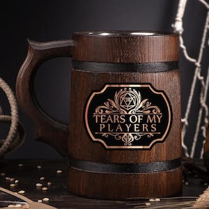 May include: A wooden beer stein with a dark brown finish and a decorative handle. The stein features a black label with the text "TEARS OF MY PLAYERS" in a copper-colored font, along with a D20 symbol and decorative flourishes. The stein is banded with black trim.