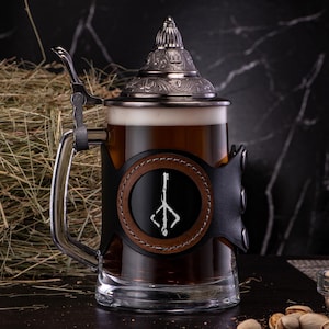 Peut inclure: Une chope en argent avec un enveloppement en cuir noir et un symbole de sorceleur. La chope est remplie de bière brune et repose sur une surface sombre.