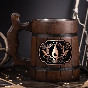 May include: A dark brown wooden beer mug with a decorative handle and a metal interior. The mug features a black and copper emblem with the word "SORCERER" and a flame design. The mug is surrounded by rope, wheat, and hops.