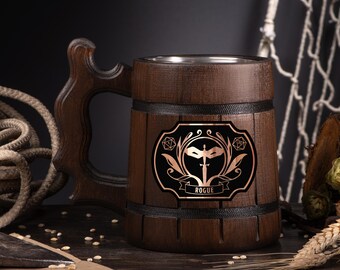 May include: A dark brown wooden beer mug with a decorative handle. The mug features a black and copper emblem with a sword, mask, and the word "ROGUE". The mug has a stainless steel interior and is banded with black trim.