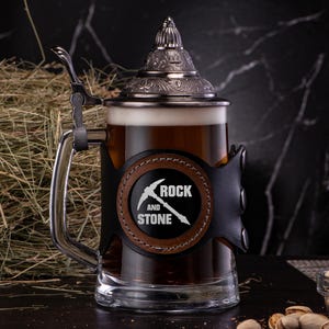 May include: A black and brown leather-wrapped beer stein with a silver lid and a metal handle. The stein has a black and brown leather patch with the text "Rock and Stone" and a pickaxe graphic.