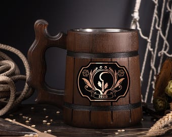 May include: A dark brown wooden beer mug with a decorative handle. The mug features a black and copper emblem with the word "Druid". The mug has a stainless steel interior and is banded with black trim. The mug is on a dark wooden surface.