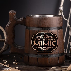 May include: A dark brown wooden beer mug with a carved handle. The mug has a metal interior and a black label with the words "DEFINITELY NOT A MIMIC" in gold lettering. The mug is decorated with black bands.
