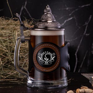 Könnte beinhalten: Ein gläserner Bierkrug mit einem Metalldeckel und einer Lederummantelung. Die Lederummantelung hat einen braunen Hintergrund mit einem weißen Design und dem Wort "Druid".