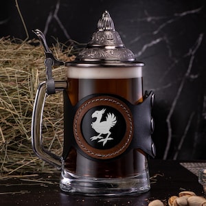 May include: A silver beer stein with a leather wrap and a white bird design. The stein is filled with dark beer and has a lid with intricate details.