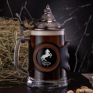 May include: A black and brown beer stein with a silver lid and a leather handle. The stein has a white logo of a horse with the text "The Prancing Pony".