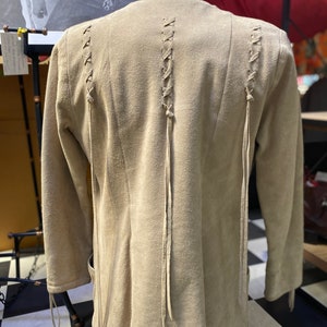 Op de afbeelding: Een beige su&egrave;de jas met lange mouwen en een vetersluiting aan de achterkant. De jas heeft een ontspannen pasvorm en een licht uitlopende zoom.