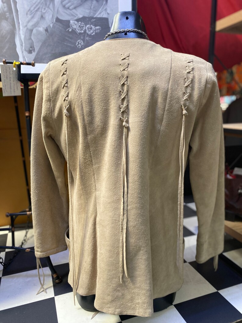 Op de afbeelding: Een beige su&egrave;de jas met lange mouwen en een vetersluiting aan de achterkant. De jas heeft een ontspannen pasvorm en een licht uitlopende zoom.