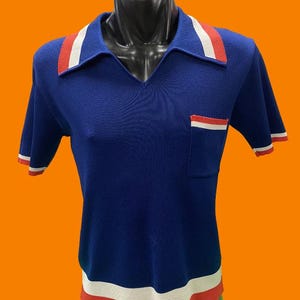 Könnte beinhalten: Ein blaues, kurzärmeliges Hemd mit roten und weißen Biesen am Kragen und Saum. Das Hemd hat eine kleine Tasche auf der linken Seite mit roten und weißen Biesen.