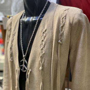 Op de afbeelding: Een beige su&egrave;de jas met lange mouwen en een V-hals. De jas heeft een vetersluiting aan de voorkant en franjes. Een zilveren ketting met een vredesduifhanger wordt over de jas gedragen.