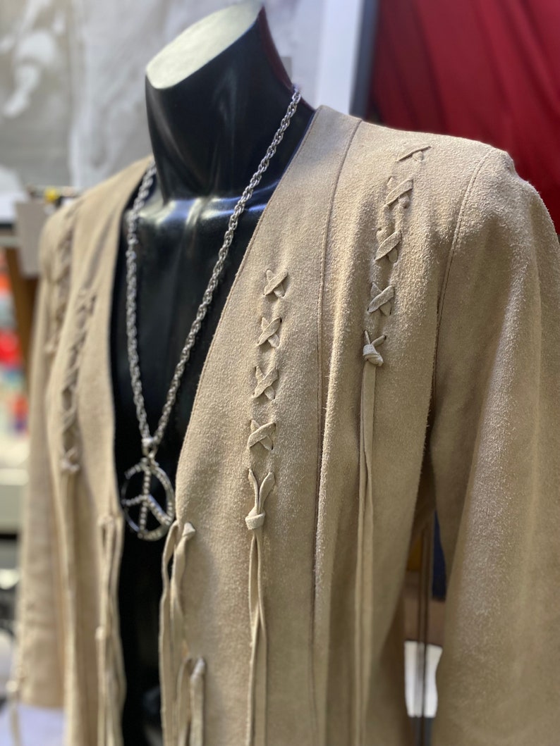 Op de afbeelding: Een beige su&egrave;de jas met lange mouwen en een V-hals. De jas heeft een vetersluiting aan de voorkant en franjes. Een zilveren ketting met een vredesduifhanger wordt over de jas gedragen.