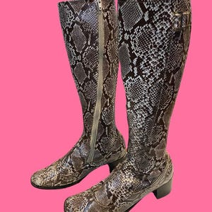 May include: Knee-high boots with a snakeskin pattern in brown, grey, and white. One boot features a zip closure, the other a buckle. The heels are approximately 5 cm high. The boots are set against a pink background.