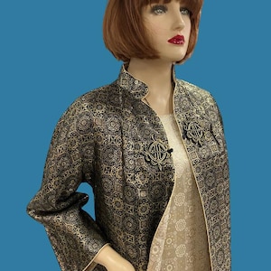 Könnte beinhalten: Eine lange Jacke aus schwarzem und goldenem Brokat mit einem Mandarinkragen und einem dekorativen Knopfverschluss. Die Jacke hat ein geometrisches Muster und ist mit einem beigefarbenen Stoff gefüttert.