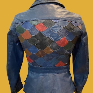 May include: A blue leather jacket featuring a back panel constructed from overlapping, multi-colored leather scales in shades of red, green, brown, black, and blue. The jacket includes a pointed collar, long sleeves, and a fitted waistline design.