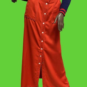 May include: A long, button-down, red skirt with white stitching and two front pockets. The skirt is paired with a navy blue sweater with red and white striped accents on the sleeves. The skirt is a vintage style.