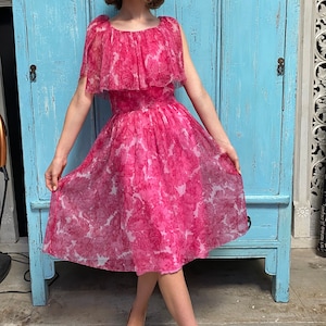Puede incluir: Un vestido rosa con estampado floral y un escote con volantes. El vestido est&aacute; hecho de una tela transparente y tiene una falda completa. La modelo lleva zapatos dorados.
