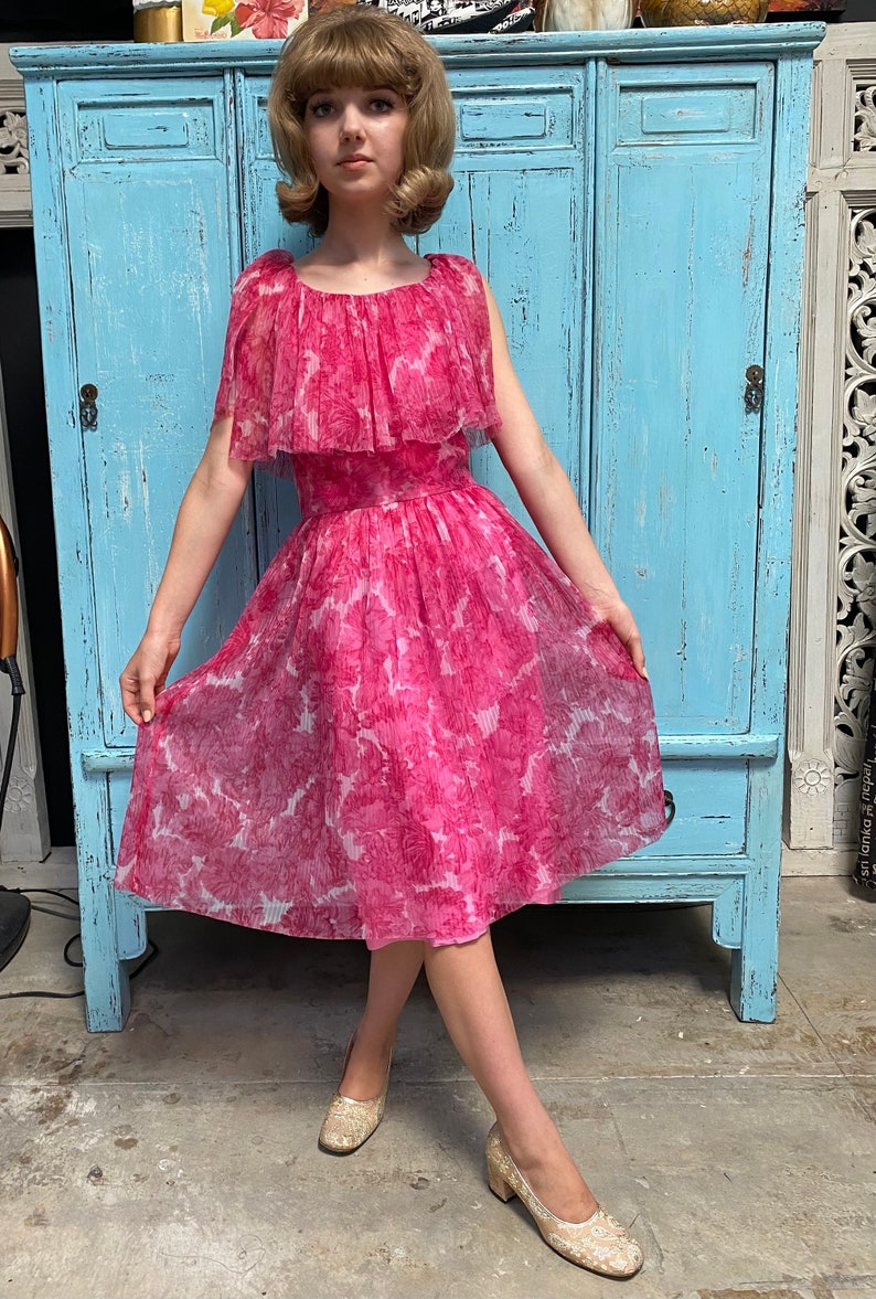 Puede incluir: Un vestido rosa con estampado floral y un escote con volantes. El vestido est&aacute; hecho de una tela transparente y tiene una falda completa. La modelo lleva zapatos dorados.