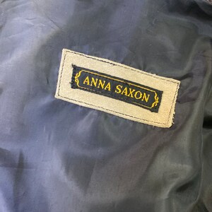 Op de afbeelding: Een close-up van een beige su&egrave;de label met zwarte stiksels en de tekst "ANNA SAXON" in gouden letters.