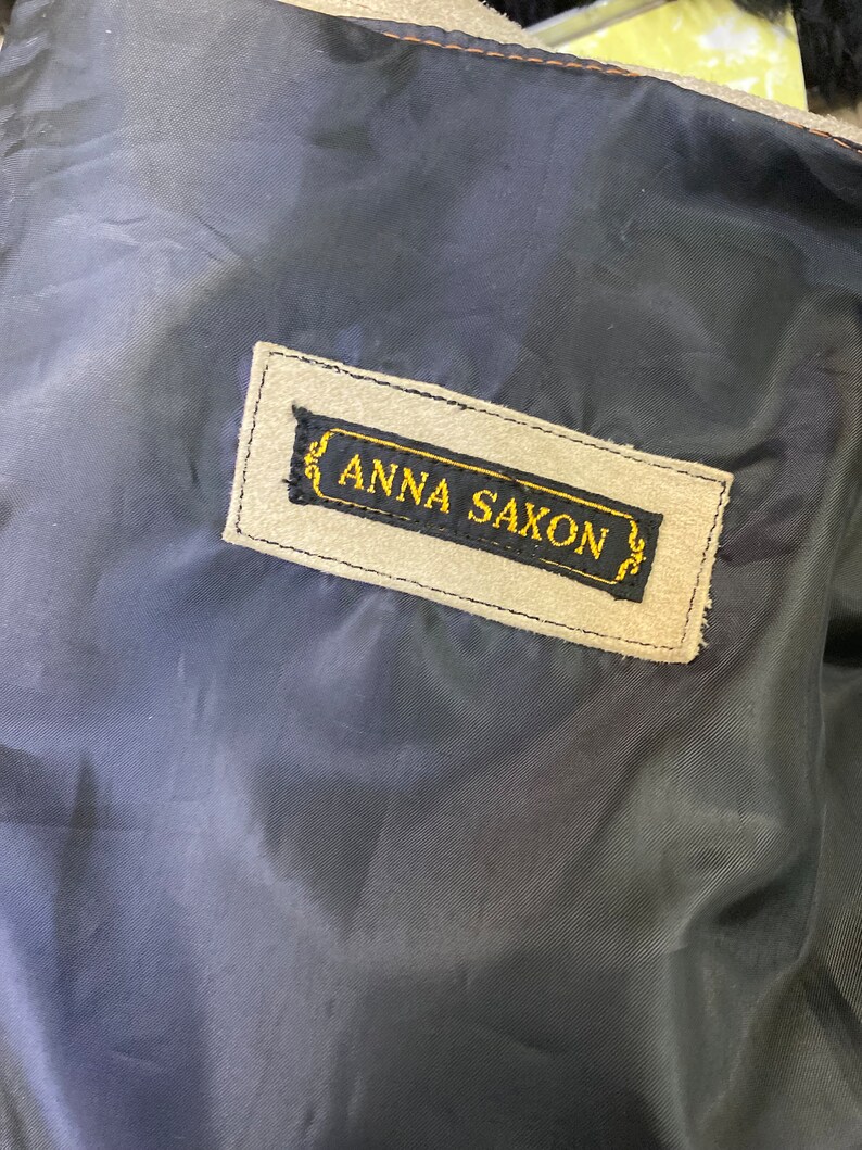 Op de afbeelding: Een close-up van een beige su&egrave;de label met zwarte stiksels en de tekst "ANNA SAXON" in gouden letters.