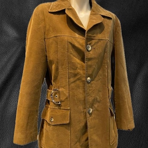 May include: A tan suede jacket with a notched collar, button closure, and two front pockets. The jacket features a belt detail at the waist and is displayed on a mannequin. The jacket has a vintage style.