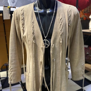 Op de afbeelding: Een beige su&egrave;de jas met franjes en een veterontwerp aan de voorkant en mouwen. De jas heeft een V-hals en wordt gedragen op een mannequin met een zilveren kettingketting en een vredesduifhanger.