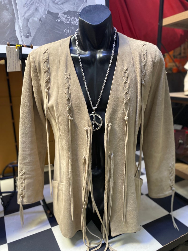 Op de afbeelding: Een beige su&egrave;de jas met franjes en een veterontwerp aan de voorkant en mouwen. De jas heeft een V-hals en wordt gedragen op een mannequin met een zilveren kettingketting en een vredesduifhanger.