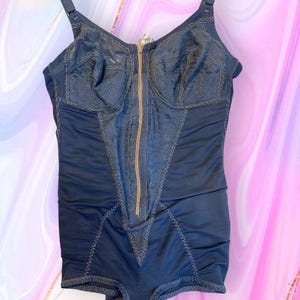 Puede incluir: Un body shaper negro con cierre de cremallera frontal y tirantes ajustables. El shaper tiene un ribete de encaje y está hecho de una tela elástica.