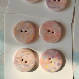 May include: Six round, decorative buttons with two holes each, arranged on white cards. The buttons feature a marbled design in pastel colors like pink, purple, yellow, and blue. The buttons are likely for sewing or craft projects.