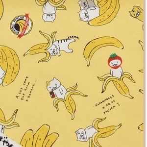Könnte beinhalten: Gelber Baumwollstoff mit einem sich wiederholenden Muster aus Katzen und Bananen. Der Stoff hat den Text "100% Fresh Banana Parlor" und "A cat came out from a banana..." aufgedruckt. Der Stoff hat auch den Text "Cotton" und "Made in Japan" aufgedruckt.