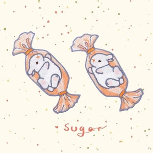 May include: Two embroidered iron-on patches featuring a white bunny inside a candy wrapper. The candy wrapper is orange and white. The text "sugar" is written below the patches.