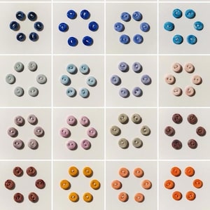 Könnte beinhalten: Ein Raster aus farbenfrohen Knöpfen, die in kreisförmigen Mustern angeordnet sind. Die Knöpfe sind in verschiedenen Farbtönen gehalten, darunter Grün, Blau, Lila, Rosa, Orange und Braun. Jeder Knopf hat zwei oder vier Löcher. Die Knöpfe befinden sich auf weißem Hintergrund.