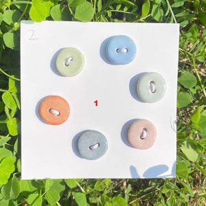 May include: Six ceramic buttons in pastel shades of orange, blue, green, and pink. Each button has two holes and a white thread. The buttons are arranged on a white surface. Numbers 1 and 2 are visible on the surface.