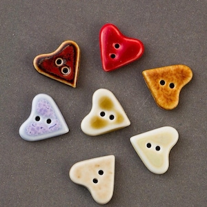 May include: A collection of heart-shaped ceramic buttons in various colors, including red, brown, and light blue. Each button has two holes for sewing. The buttons are arranged on a gray surface.