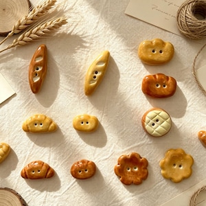 May include: A collection of twelve ceramic buttons shaped like various types of bread, including baguettes, croissants, and round rolls. Each button features a glossy, golden-brown finish and two small holes in the centre for sewing attachment.