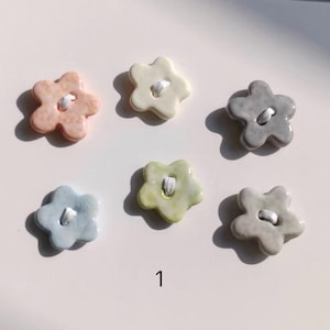May include: Six flower-shaped buttons in pastel colors including peach, cream, gray, light blue, pale green, and mottled white. Each button features a central hole with white thread visible. The number 1 is printed in black below the buttons.